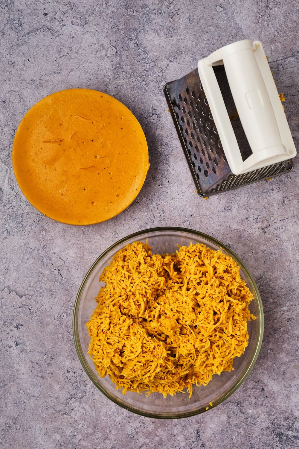 A top-down view of a box grater, a block of vegan cheese, and shredded vegan cheese in a bowl.