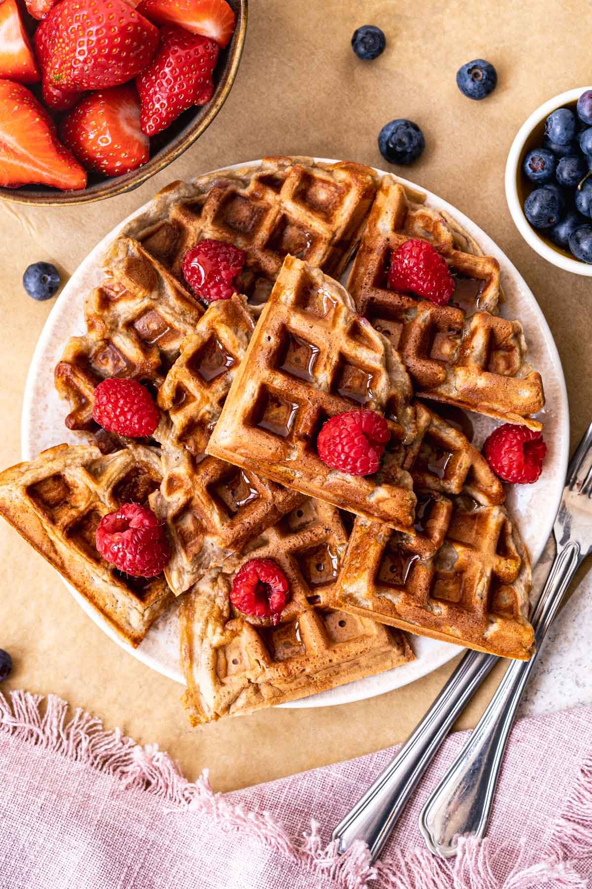 An overhead view of vegan protein waffles on a plate topped with raspberries, next to bowls of blueberries and strawberries. 