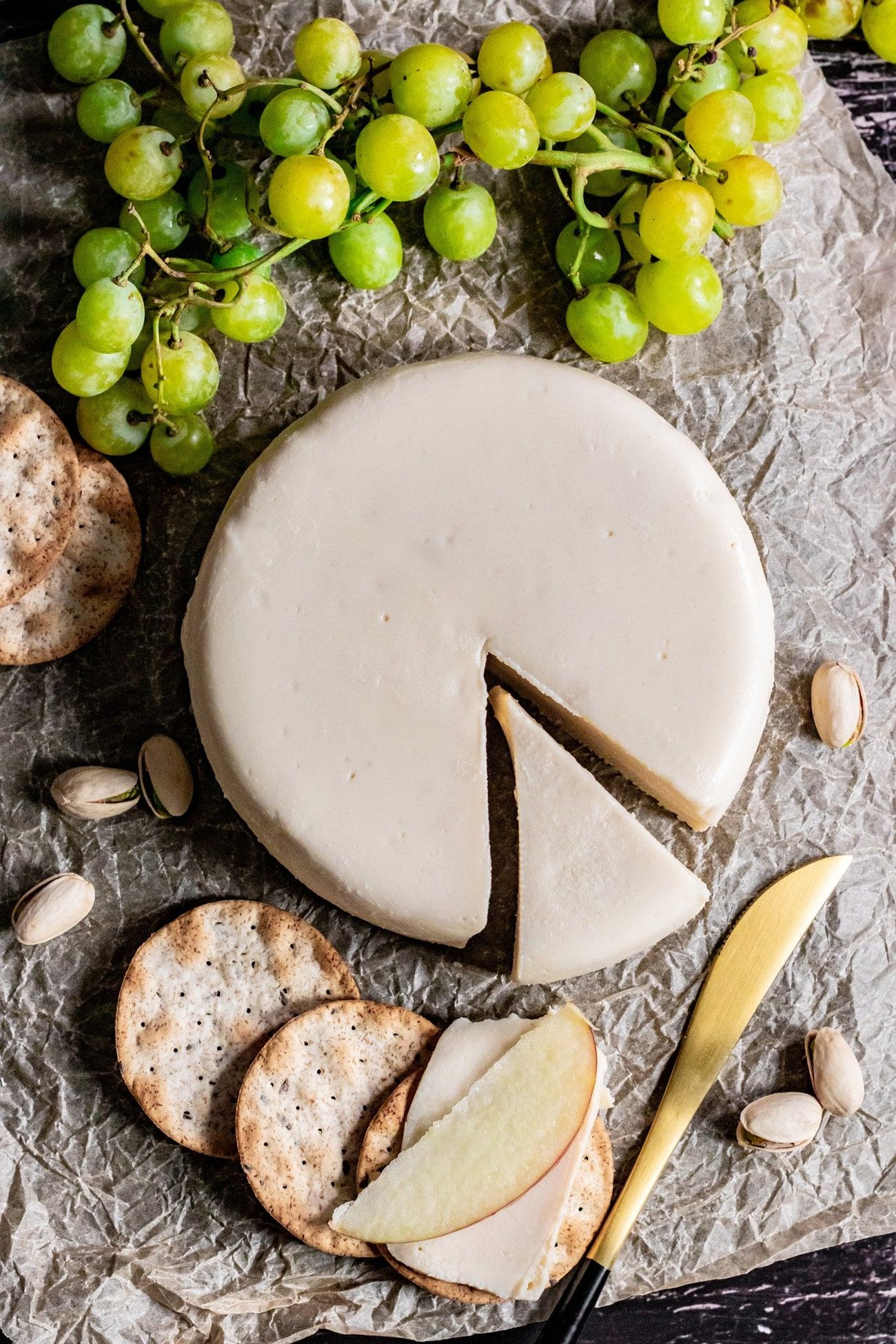 Vegan brie on a table, surrounded by grapes and crackers with an apple slice. 