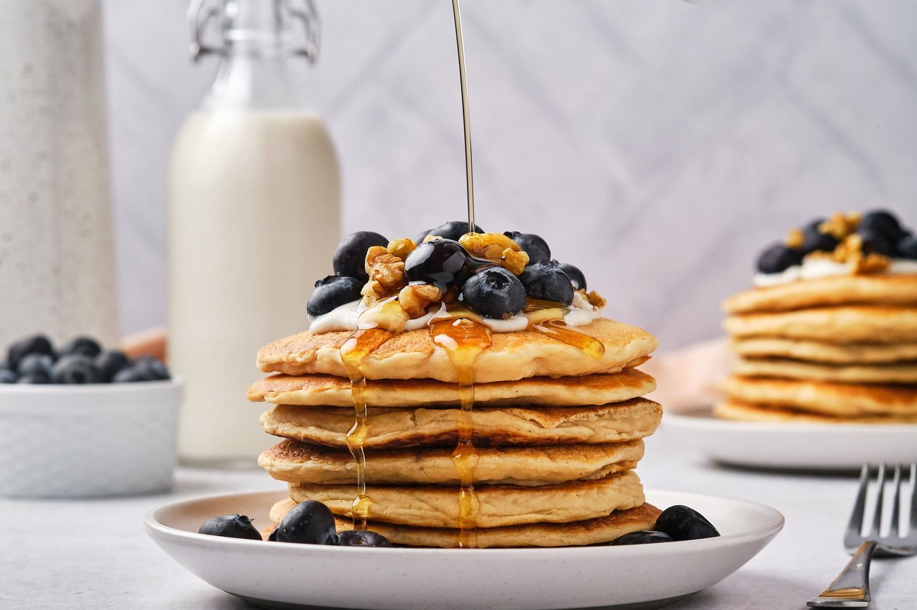 A stack of vegan protein pancakes on a plate with maple syrup being poured on top.