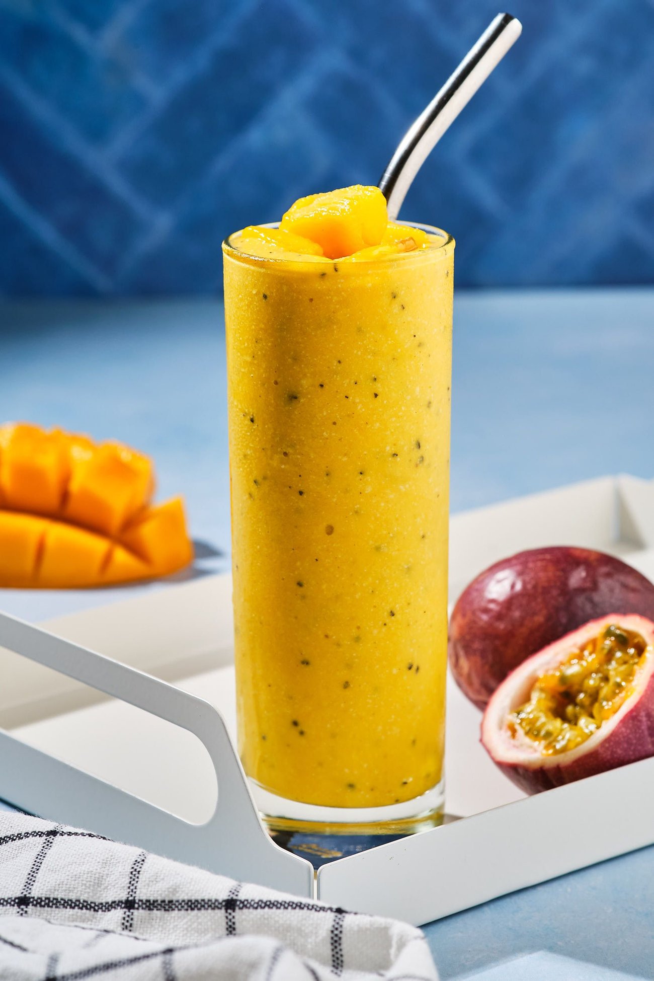 A mango smoothie on a table with a straw, surrounded by fresh cut fruits and linens.