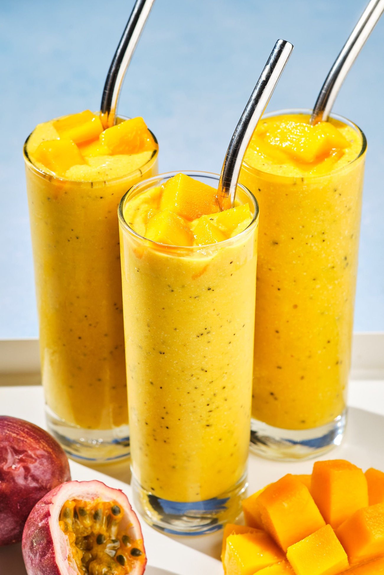 Three mango smoothies on a drink tray with cut mango and passion fruit.