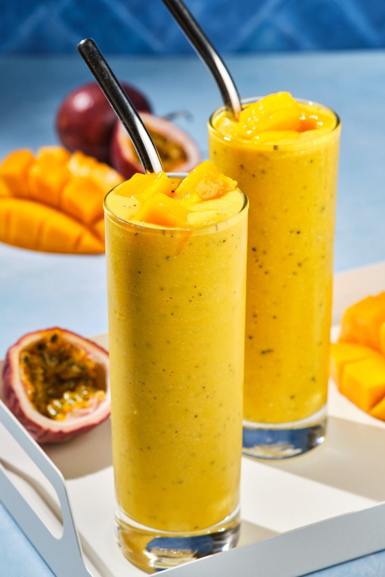 Two mango turmeric smoothies on a drink tray with fresh cut fruits in the background.