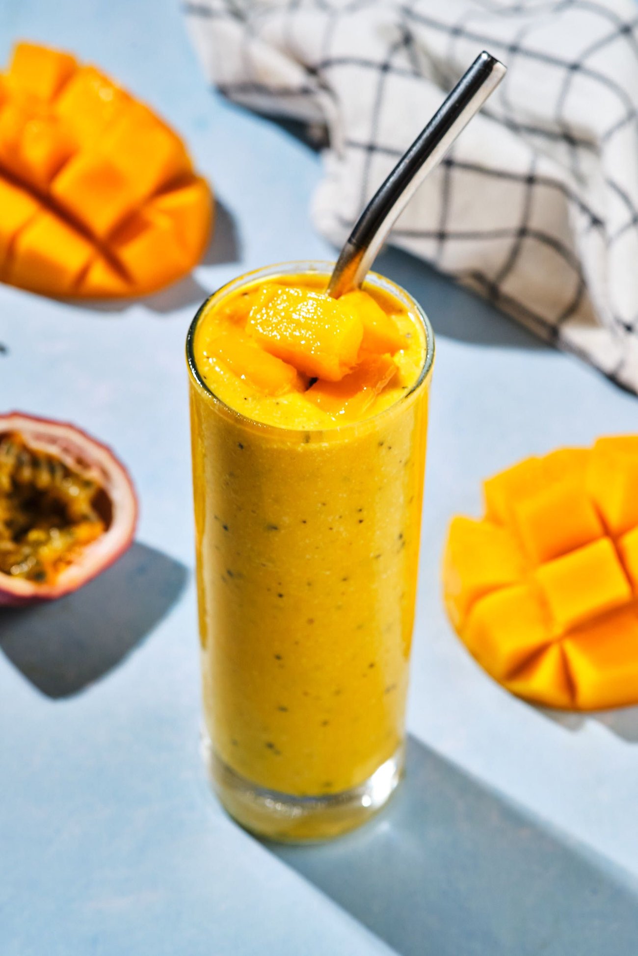 A mango smoothie in a glass with a straw, topped with fresh mango chunks, on a table with cut mangoes and passion fruit.