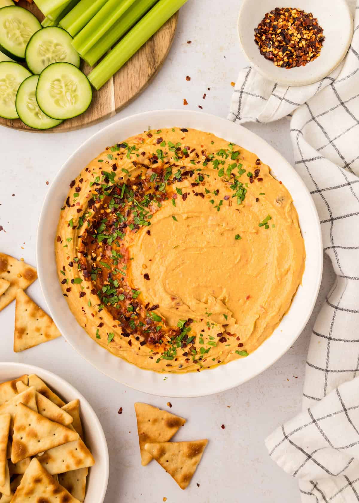 Roasted red pepper hummus in a bowl next to a bowl of pita chips and a platter of vegetables. 