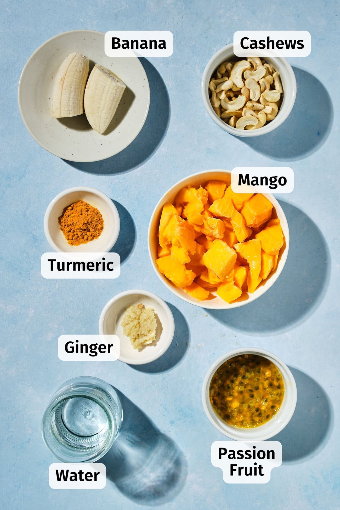 The ingredients for a mango smoothie in individual bowls on a table, labeled.