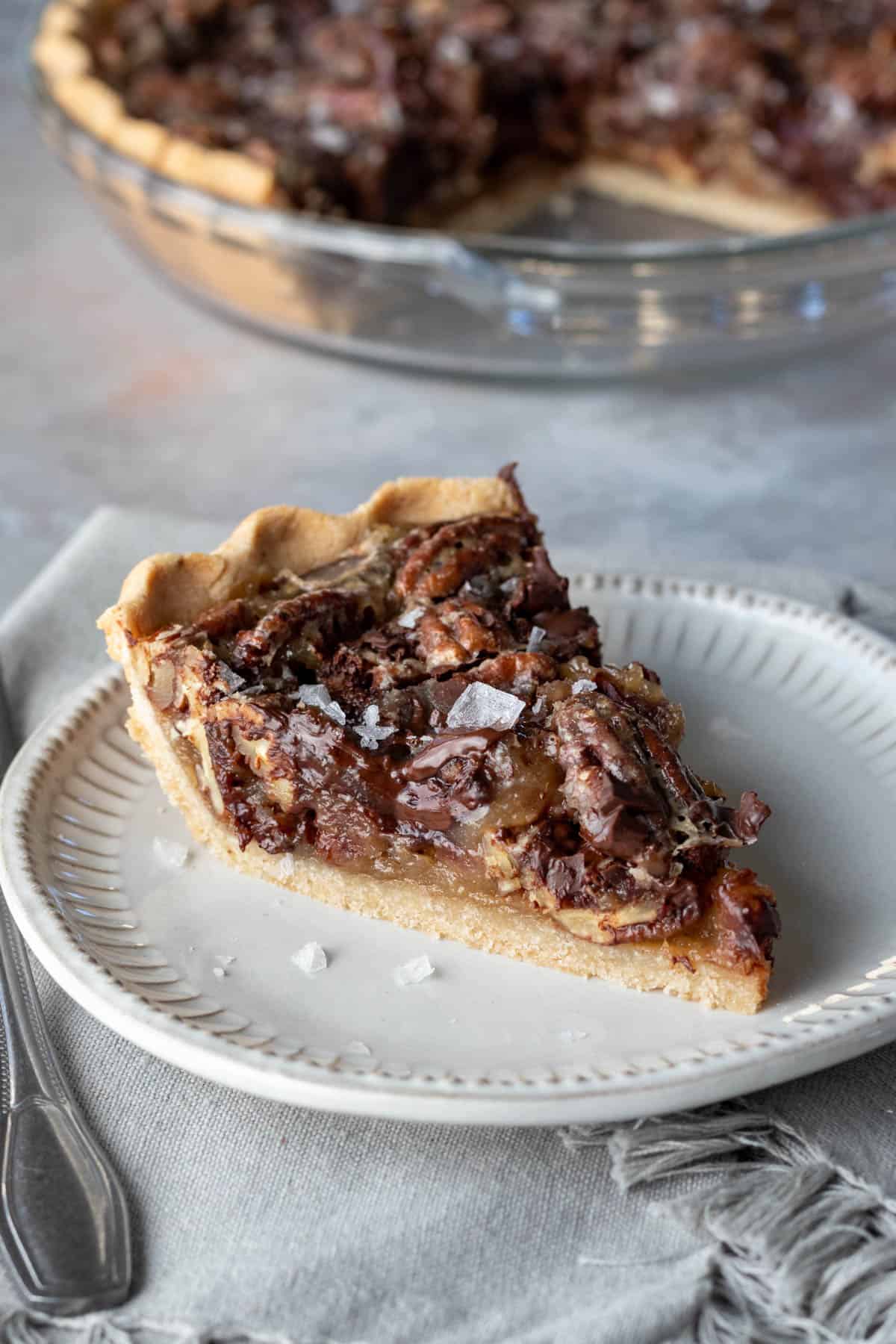 A slice of vegan salted chocolate pecan pie on a plate, with the remainder of the pie in the background. 