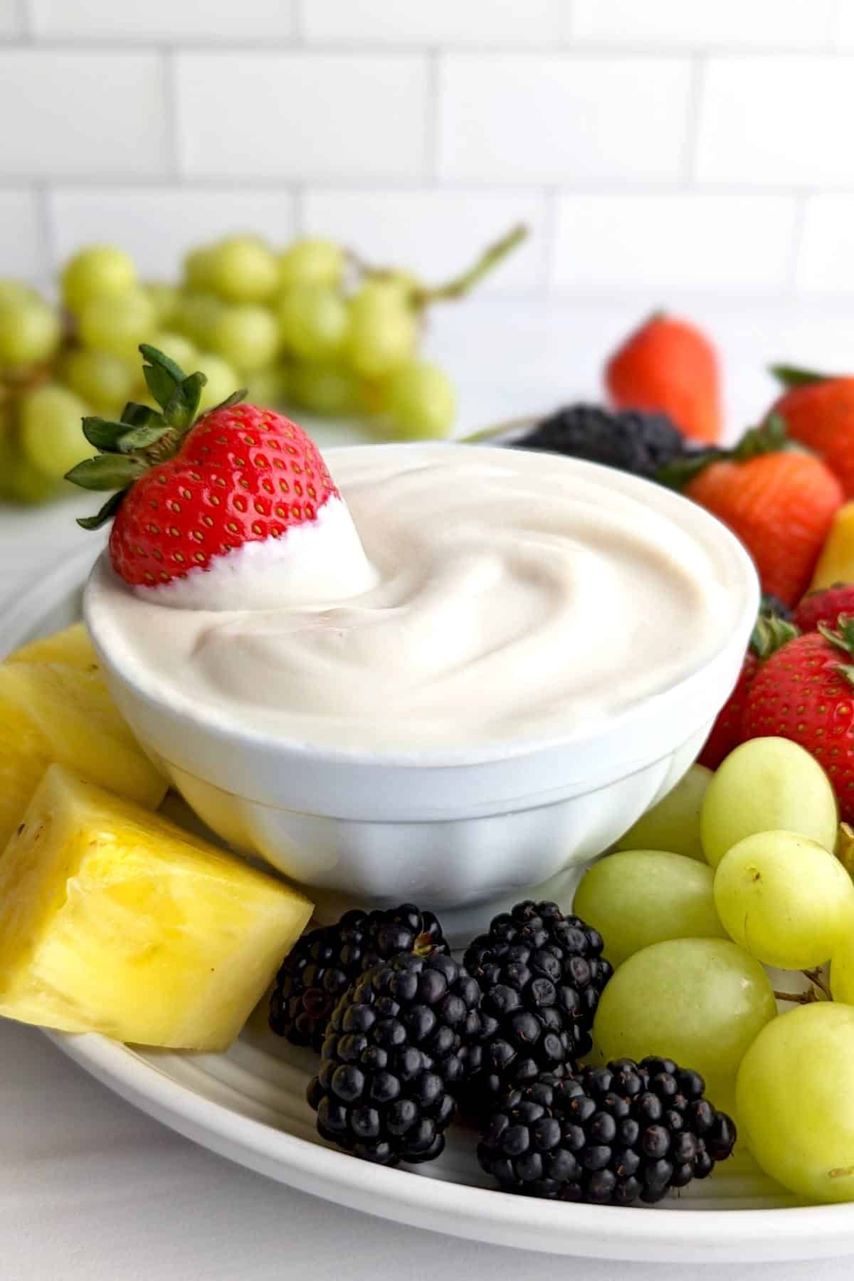 Vegan fruit dip in a bowl surrounded by a plate of fruits including strawberries, grapes, pineapple, and blackberries.