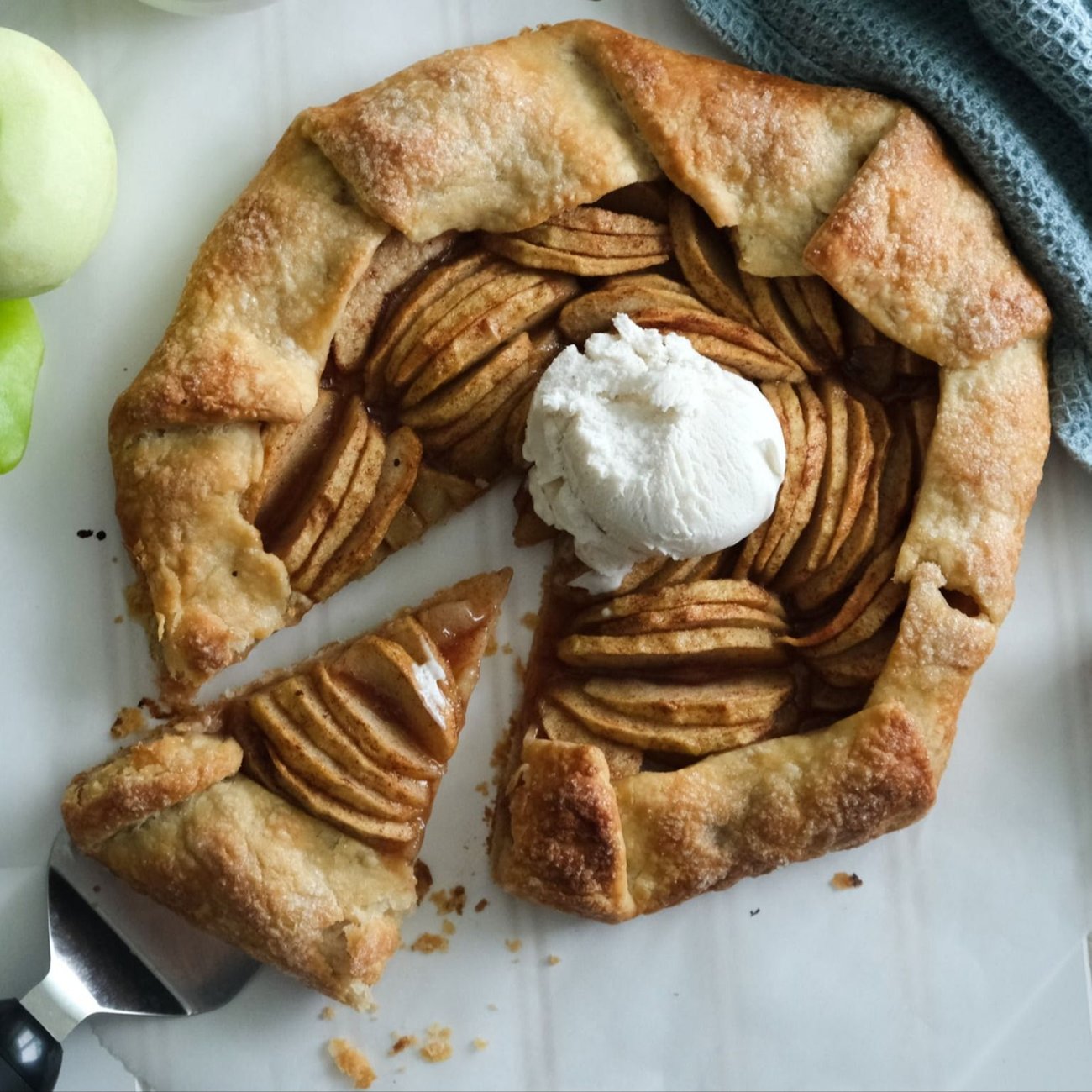 An apple galette with a slice being pulled away, topped with vegan ice cream; a peeled apple to the left, and a ramekin of brown sugar above.