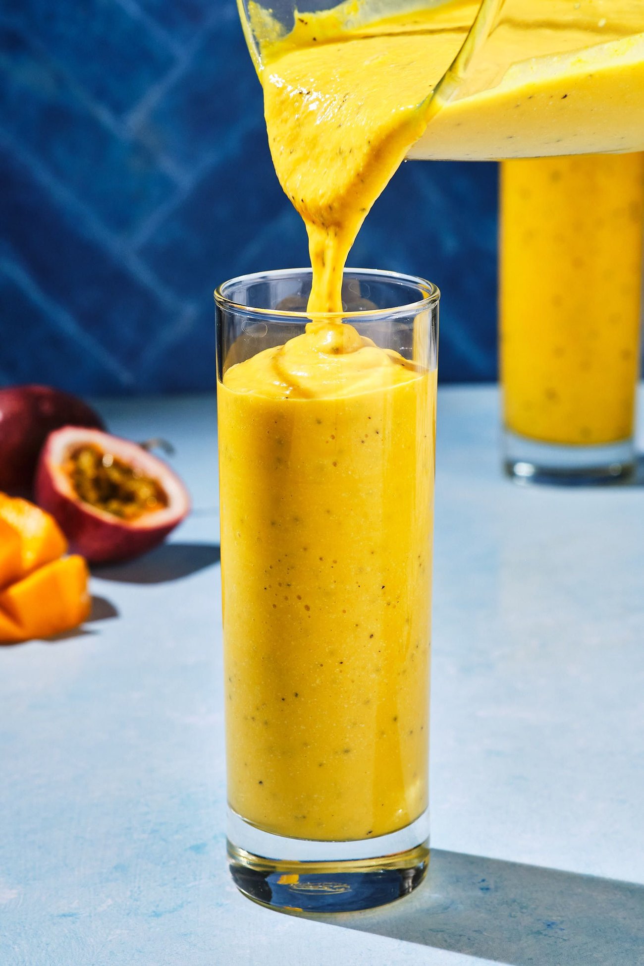 A mango smoothie being poured from a blender pitcher into a tall glass; cut mangoes and passion fruits are in the background.