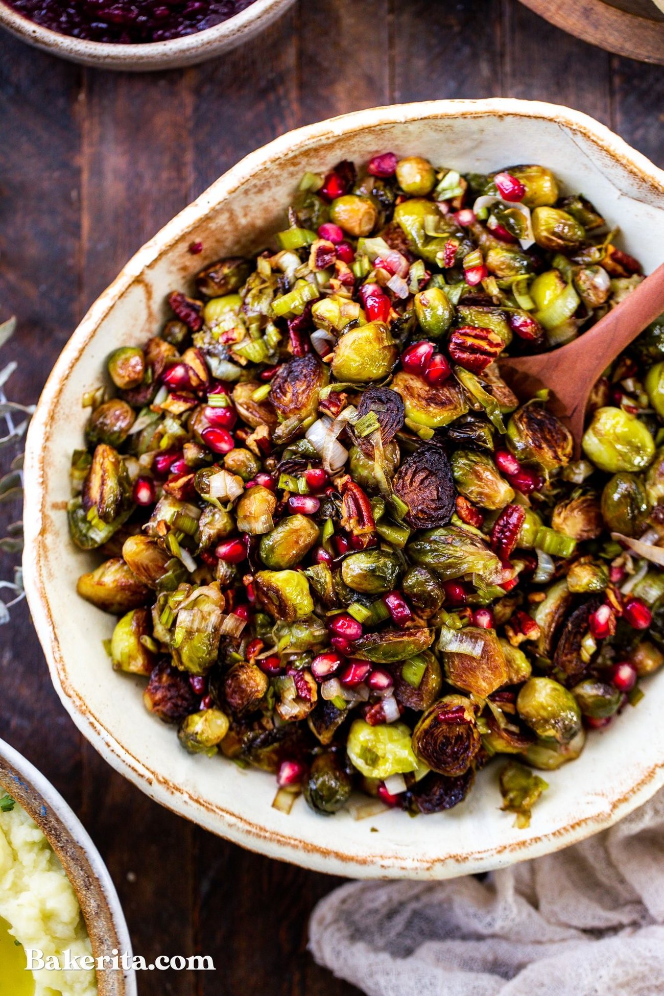 A top-down view of a large bowl of roasted brussels sprouts with pecans and pomegranate arils with a serving spoon.