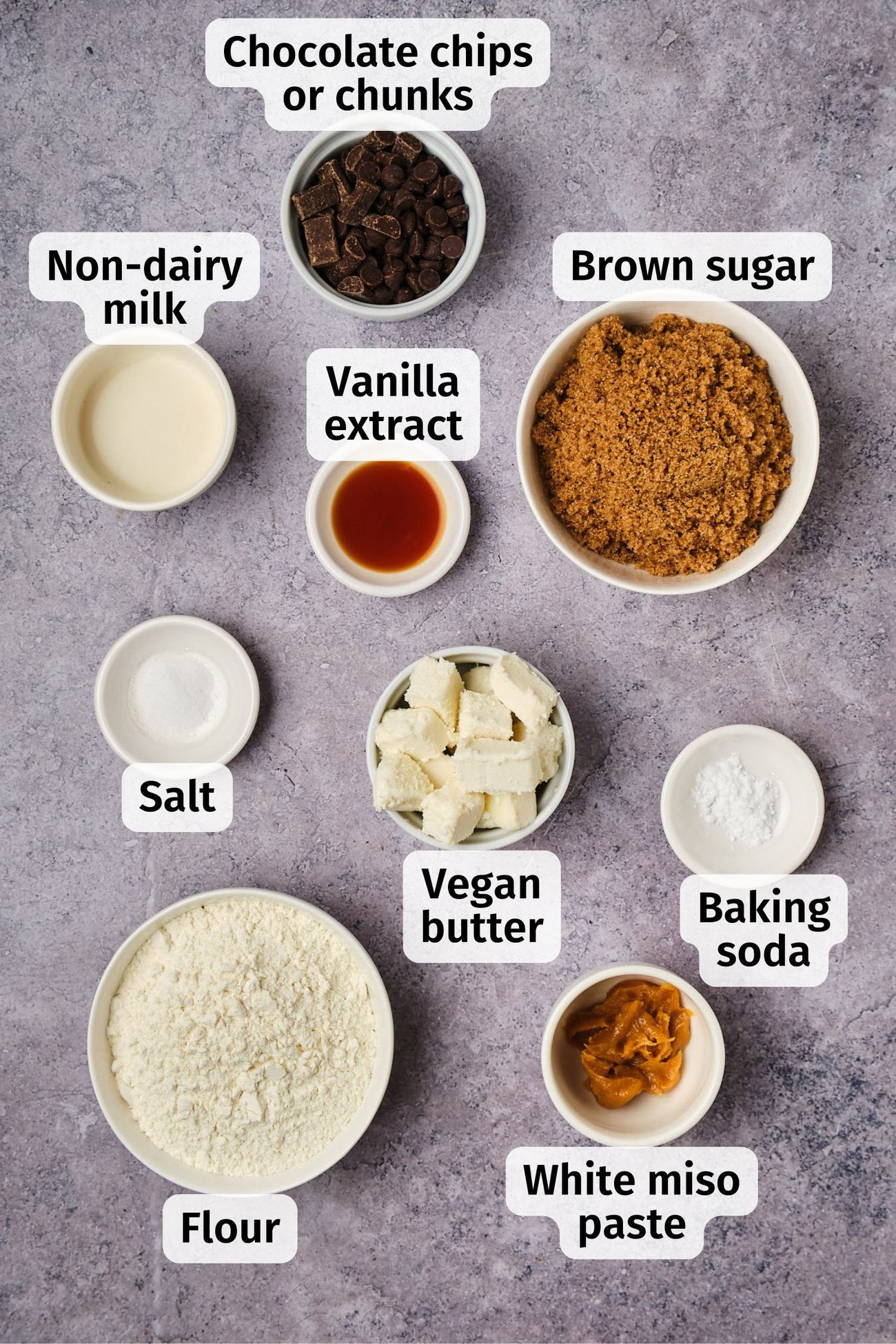 The ingredients for vegan chocolate chip cookies in bowls on a table, labeled.