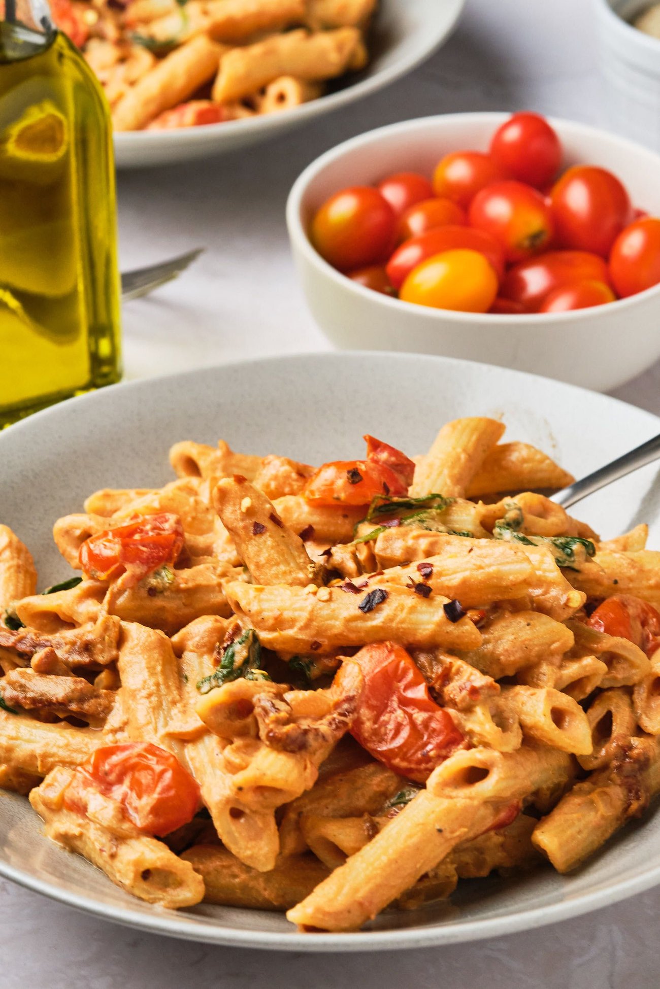 Creamy tomato pasta, served in a pasta bowl on a table next to a bottle of olive oil and a bowl of grape tomatoes.