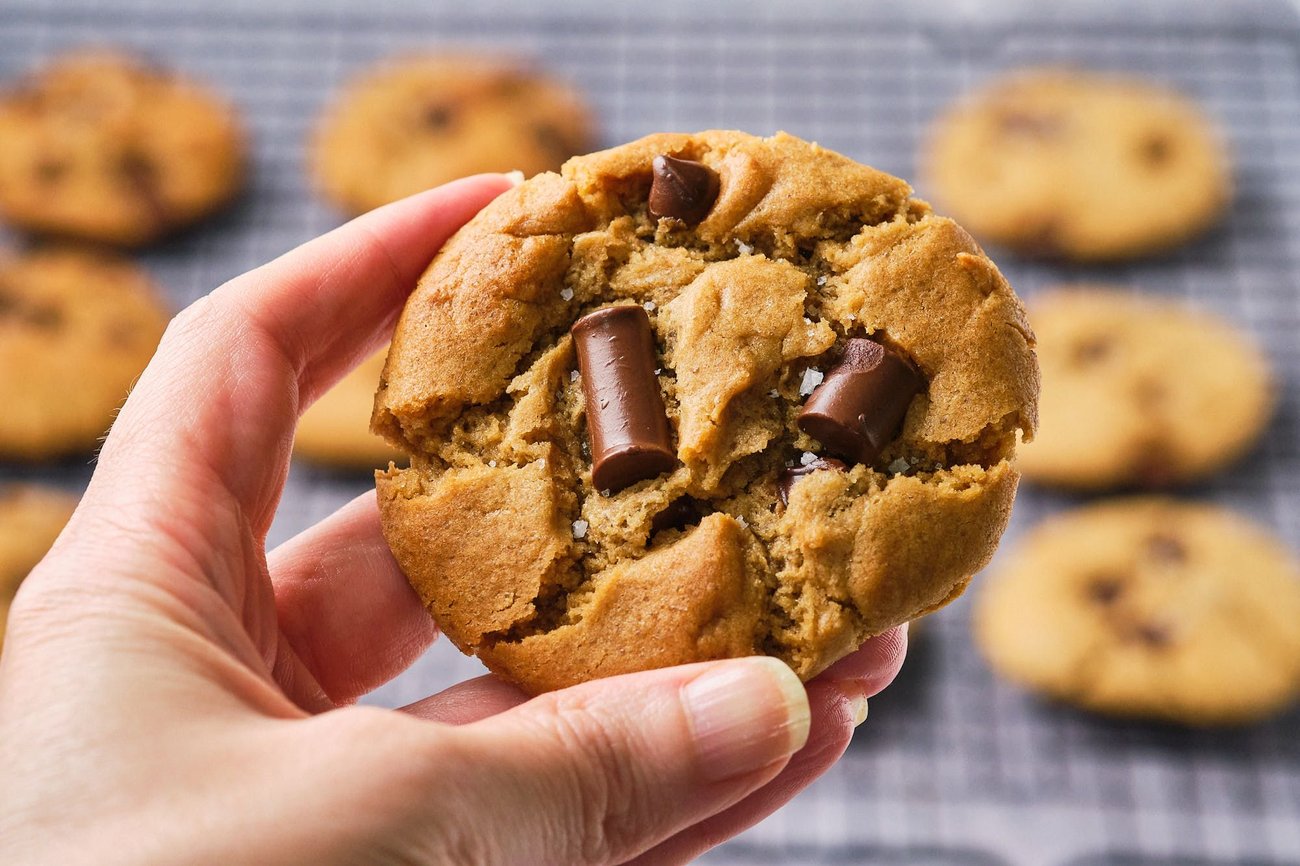 A hand holding a vegan chocolate chip cookie with cookies on a wire rack in the background.