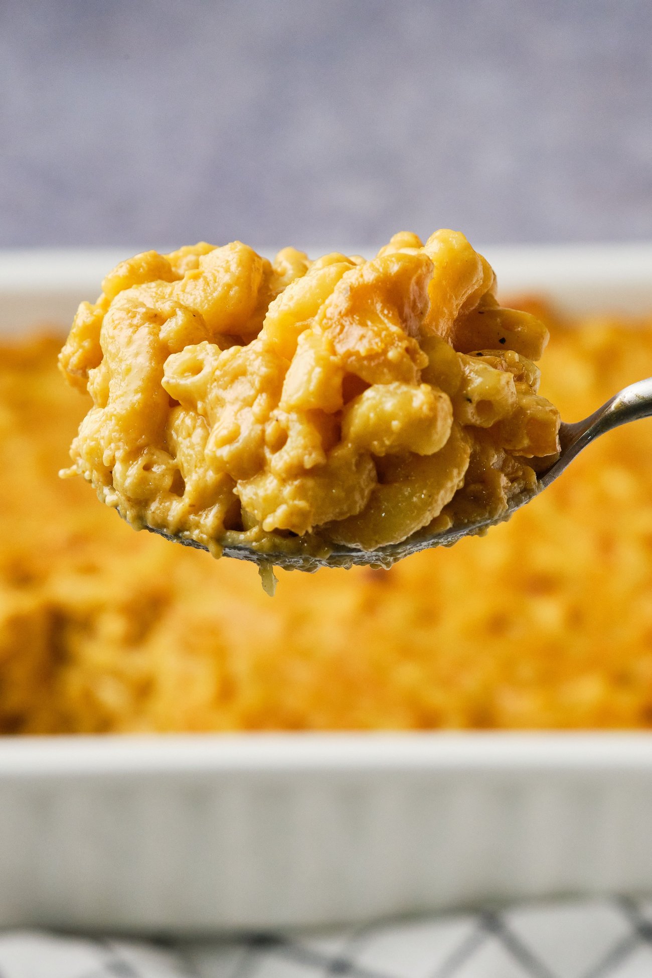 Vegan mac and cheese on a serving spoon (close up) with a casserole dish of mac and cheese in the background.