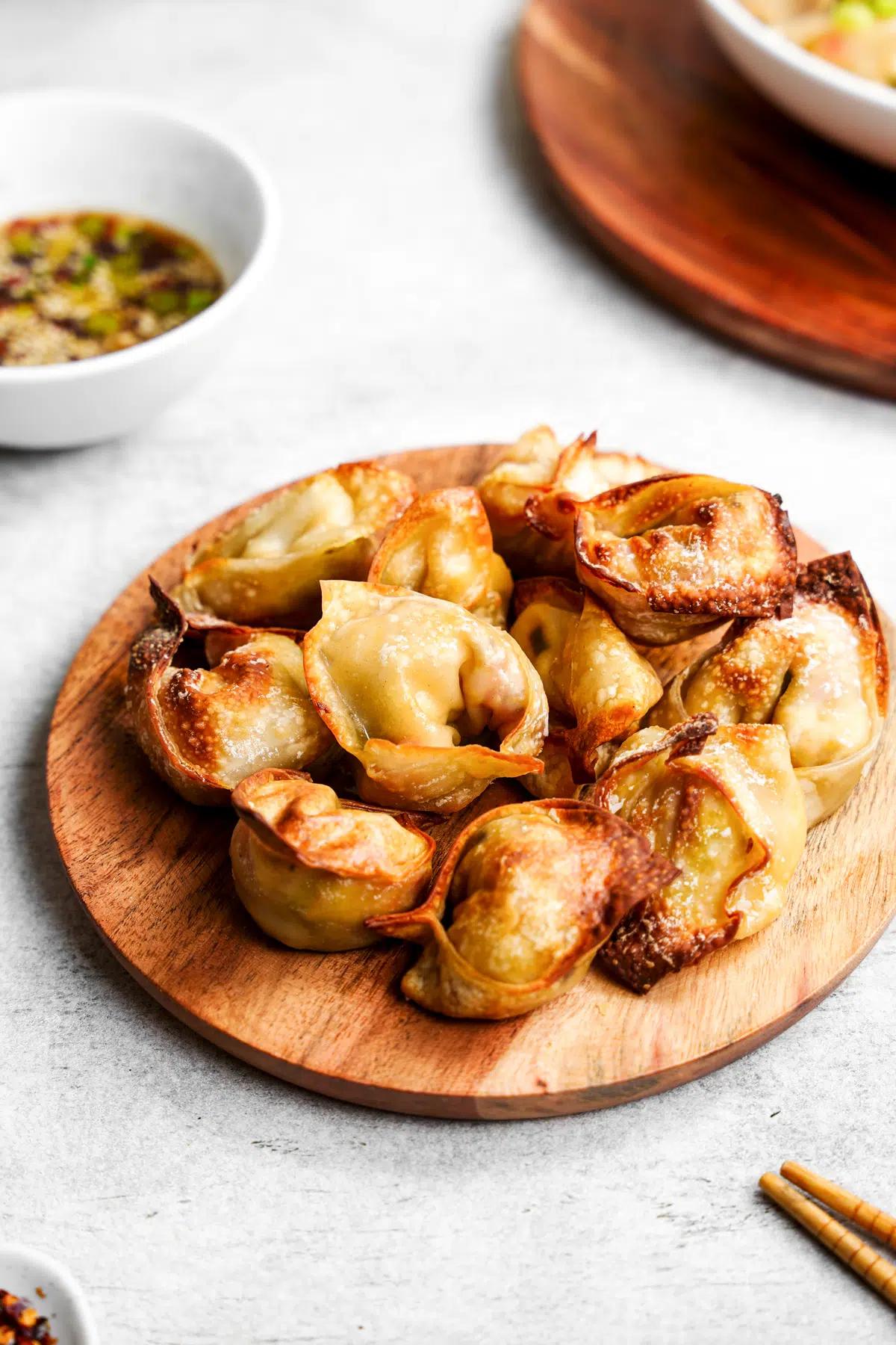 A bowl of air-fried tofu wontons on a round wooden platter with chopsticks in the foreground and a bowl of dipping sauce in the background.