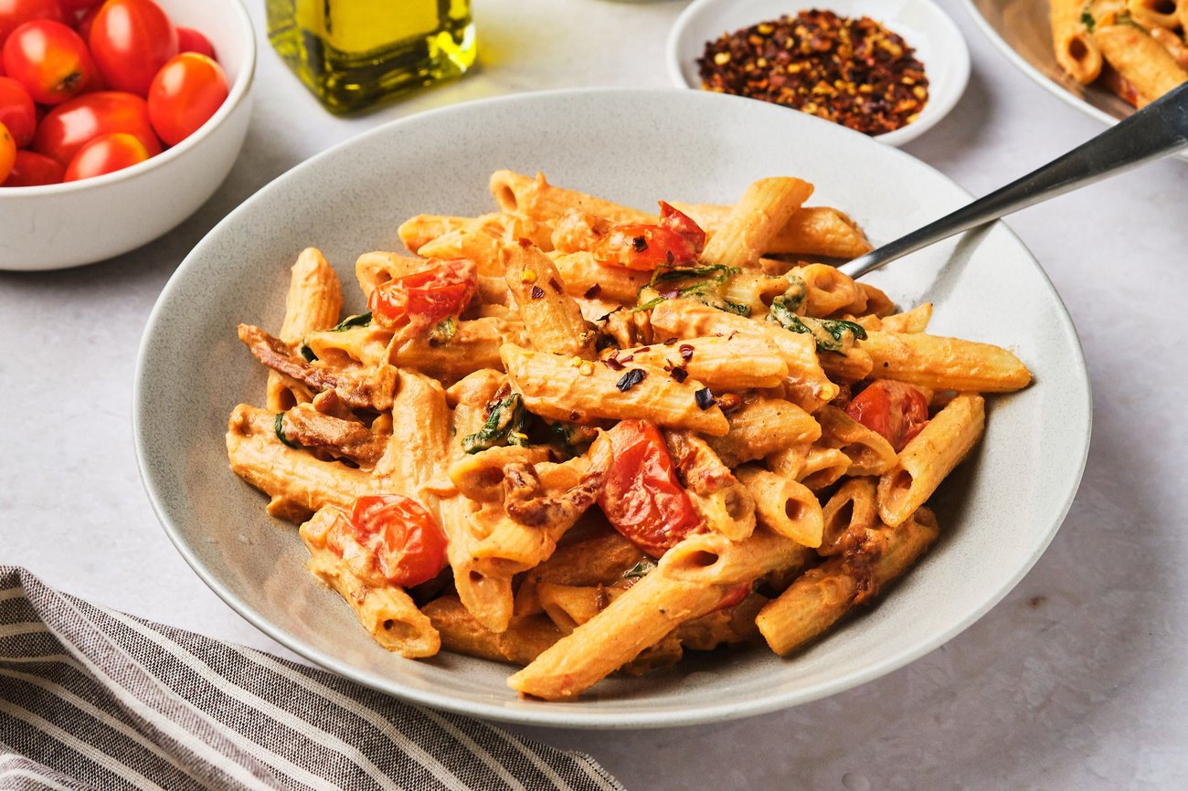 Vegan creamy tomato pasta in a bowl on a table with red pepper, olive oil, and grape tomatoes. 
