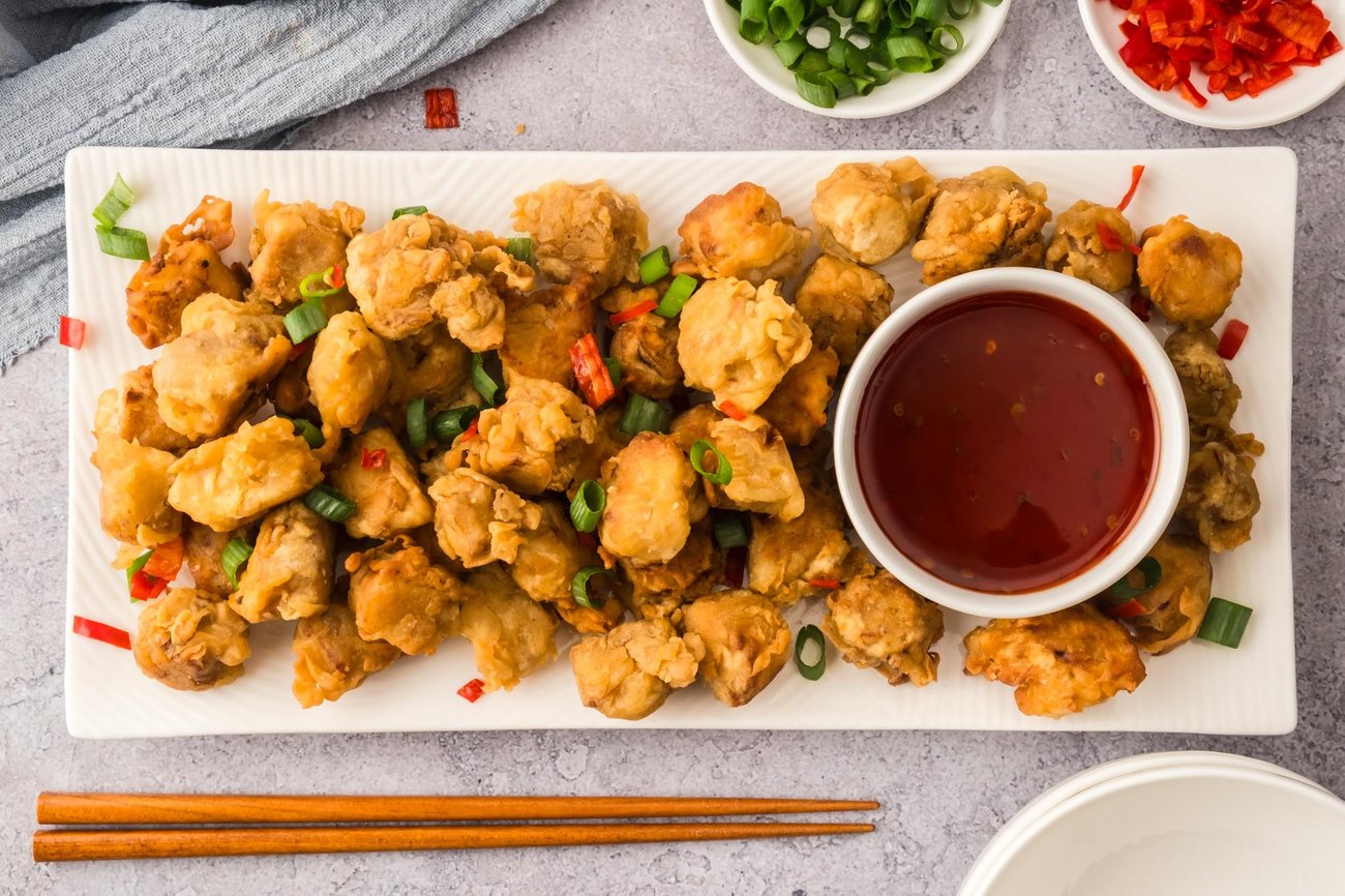 Fried tofu on a large platter with a small ramekin of sauce in the middle, next to chopsticks and small bowls of green onions and peppers.