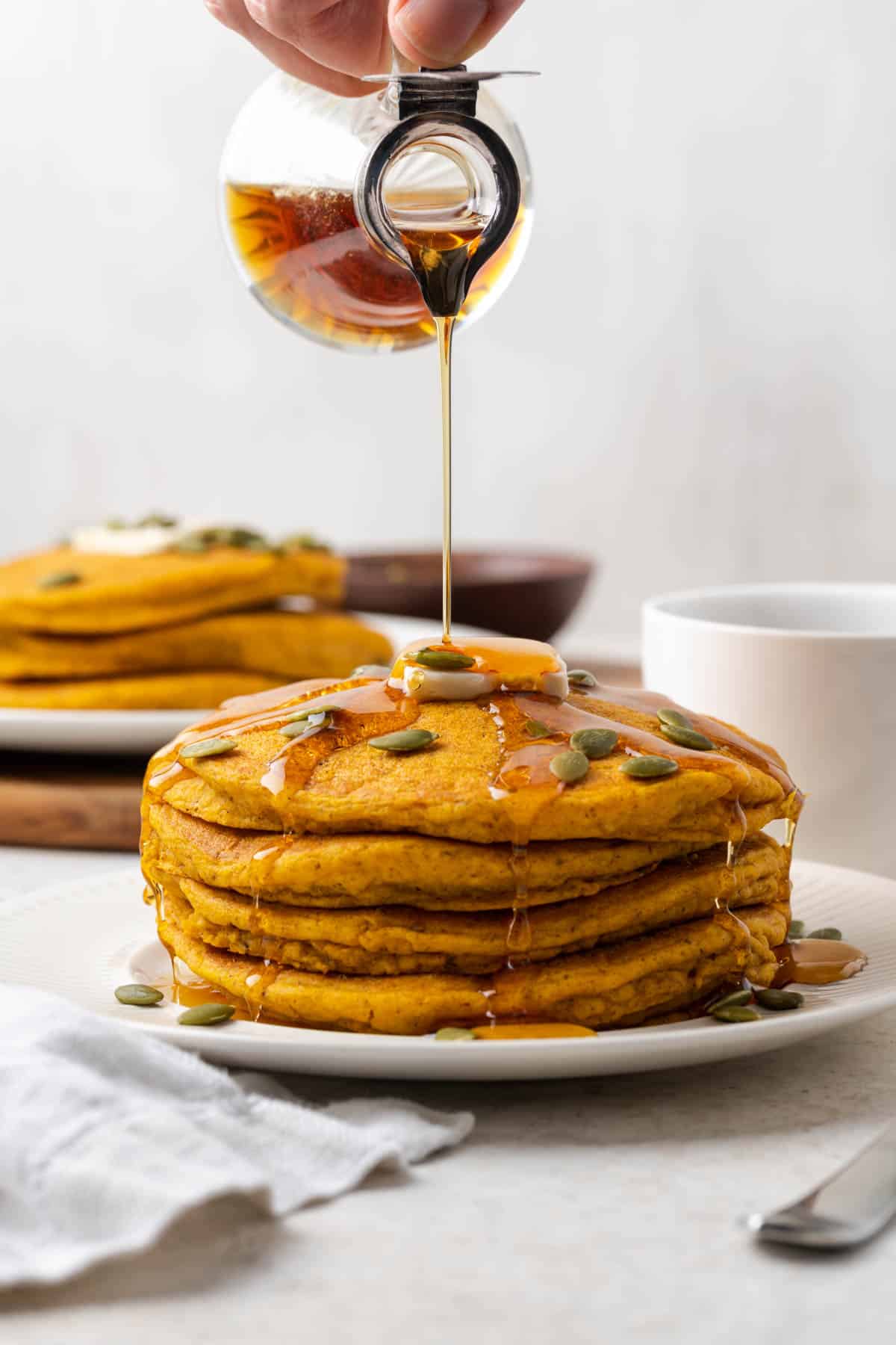 A hand pouring maple syrup over a stack of vegan pumpkin pancakes on a plate. 