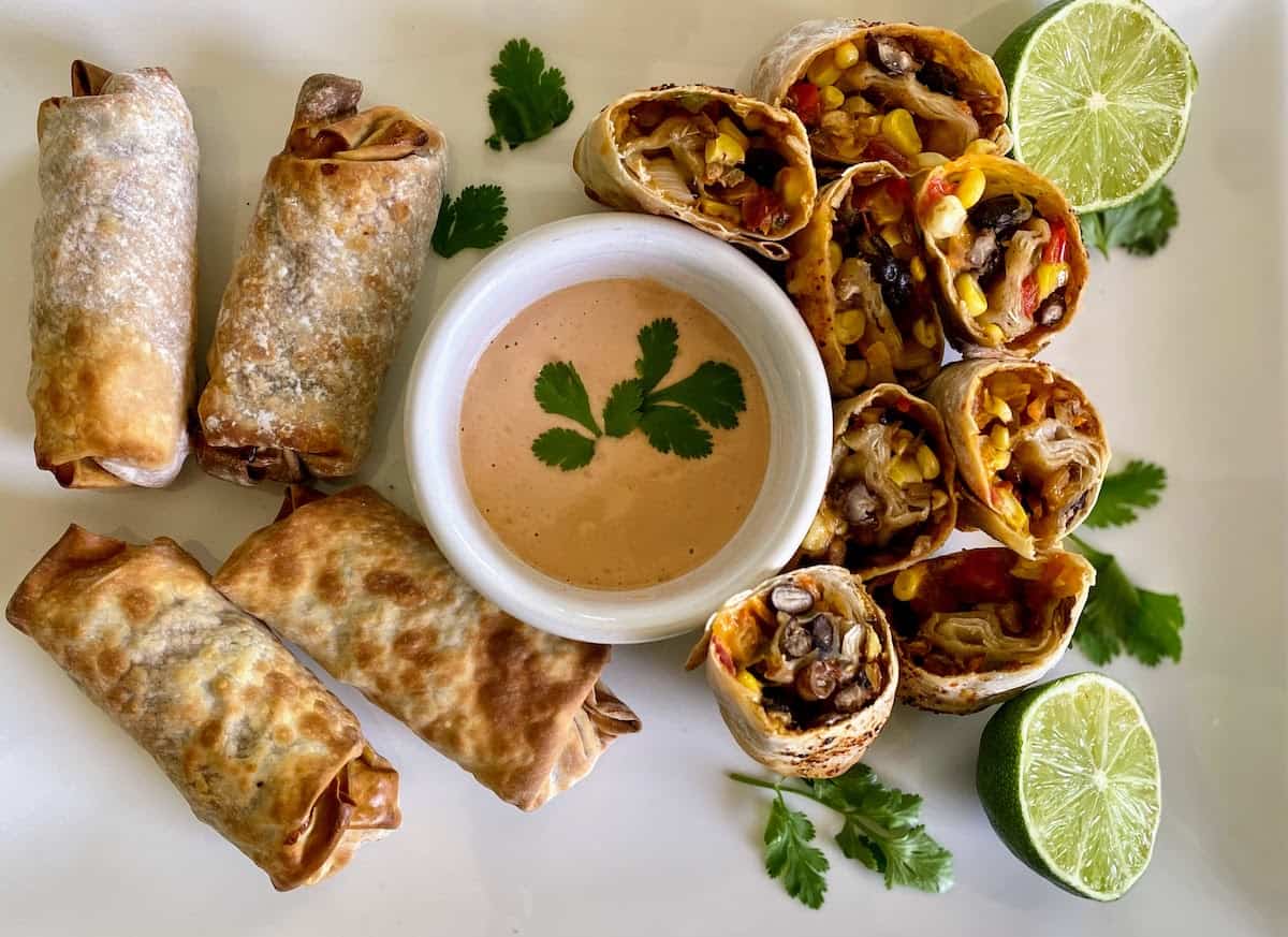 Vegan Southwest egg rolls on a table with a bowl of dipping sauce and lime halves; the egg rolls on the right side are cut to show their filling.