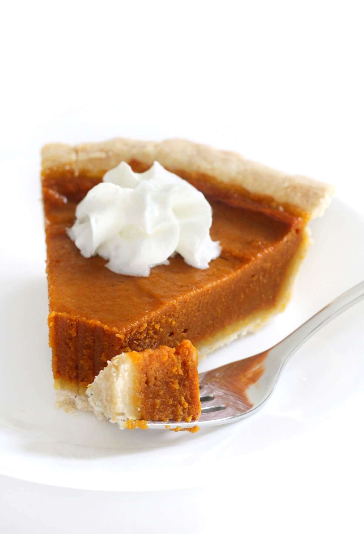 A slice of vegan and gluten-free pumpkin pie topped with vegan whipped cream, with a piece of pie on the fork next to it. 