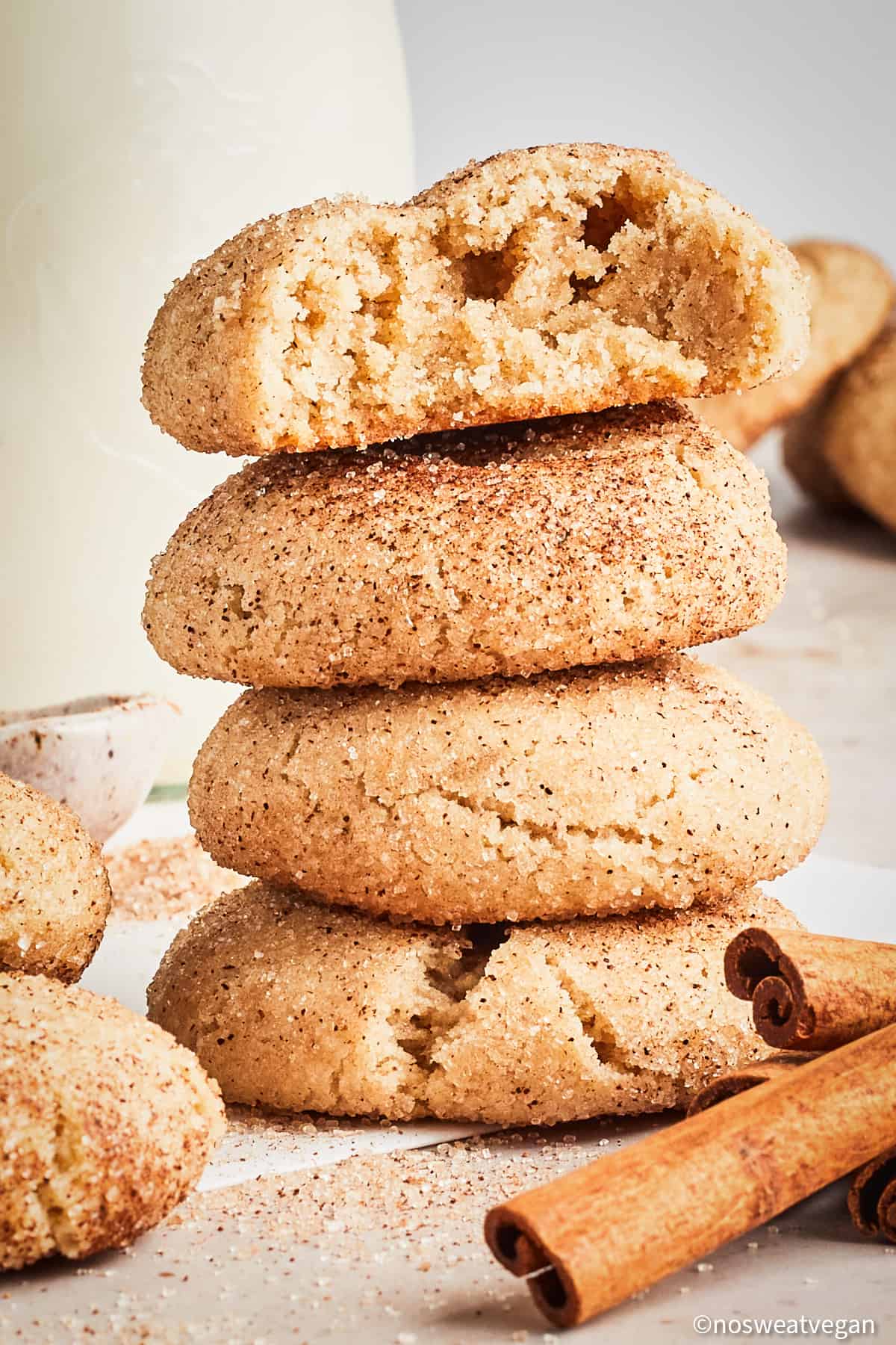 A stack of vegan snickerdoodle cookies next to cinnamon sticks and a jug of plant milk. 