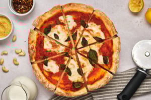 A sliced vegan pizza with a jar of liquid mozzarella to the side, as well as cashews, vegan parmesan, crushed red pepper, lemons, and a pizza cutter.