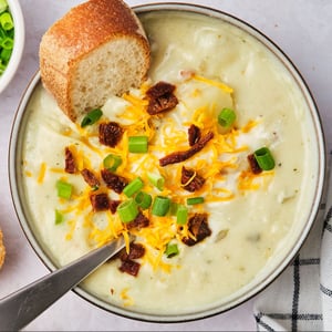 Vegan potato soup in a bowl with a slice of bread, topped with vegan sour cream, vegan cheddar cheese, sun-dried tomatoes, and green onions.