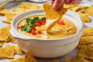 A hand dipping a tortilla chip into a pot of vegan queso dip, surrounded by tortilla chips on the table. 