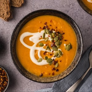 A top-down image of a bowl of creamy carrot pumpkin soup topped with cashew cream, red pepper flakes, and pepitas.