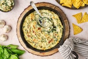 A top-down view of vegan spinach and artichoke dip in a cast-iron pan, with a spoon. To the sides are garlic, spinach, chips, and a bowl of dip.