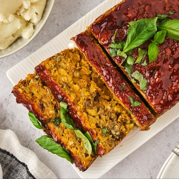 Top-down view of a lentil loaf on a platter, sliced, with plates and mashed potatoes to the sides.