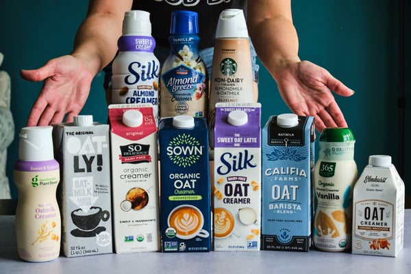 Eleven vegan creamers lined up on a table, with hands behind them.