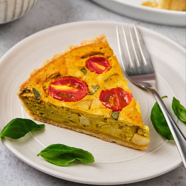 A slice of vegan quiche on a plate with a fork and basil leaves. 