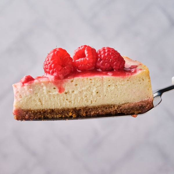 A slice of vegan cheesecake on a pie server, topped with raspberries and raspberry compote. 