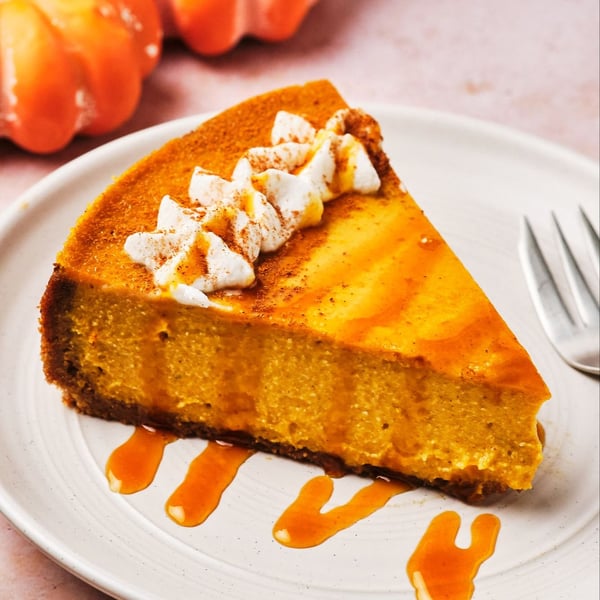 A slice of vegan pumpkin cheesecake topped with vegan whipped cream and caramel, on a plate with a fork; mini pumpkins are in the background.