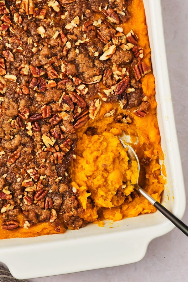 A close-up view of vegan sweet potato casserole in a casserole dish, topped with pecans and brown sugar, with a serving spoon at the corner. 