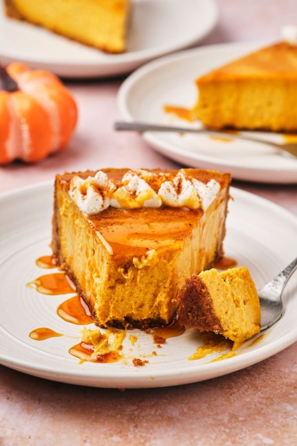 A slice of vegan pumpkin cheesecake topped with vegan whipped cream and caramel, on a plate with a fork and a piece taken out.
