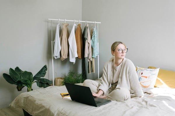 A woman working on her laptop on her bed, looking off to the side.
