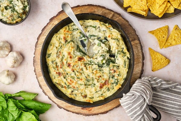 A top-down view of vegan spinach and artichoke dip in a cast-iron pan, with a spoon. To the sides are garlic, spinach, chips, and a bowl of dip.