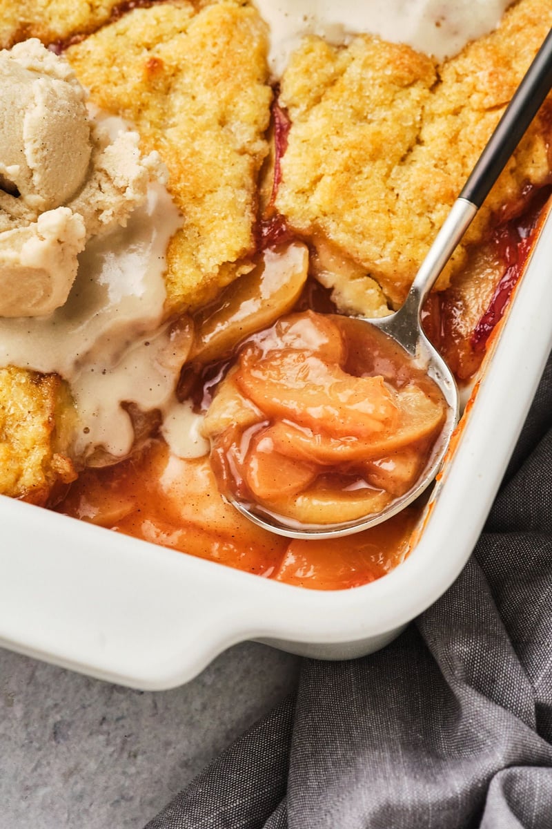 Vegan peach cobbler in a casserole dish with a serving spoon and scoops of ice cream. 