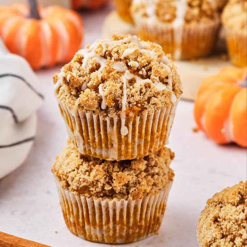 A stack of 2 vegan pumpkin muffins with more muffins and mini pumpkins in the background. 