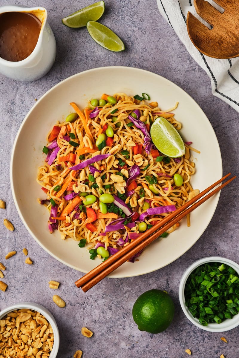 A top-down view of a Thai noodle salad in a bowl with a lime wedge and chopsticks, surrounded by bowls of green onions, peanuts, and peanut sauce.