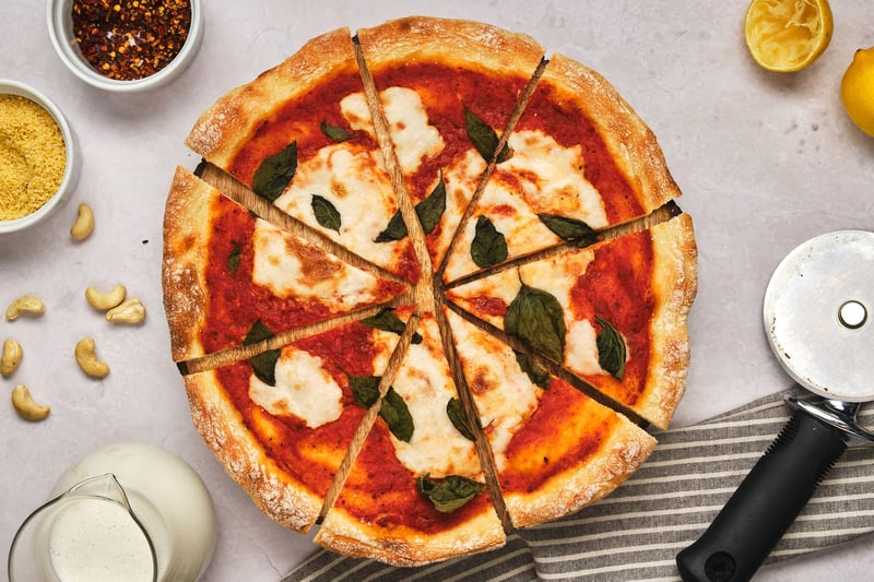 A sliced vegan pizza with a jar of liquid mozzarella to the side, as well as cashews, vegan parmesan, crushed red pepper, lemons, and a pizza cutter.