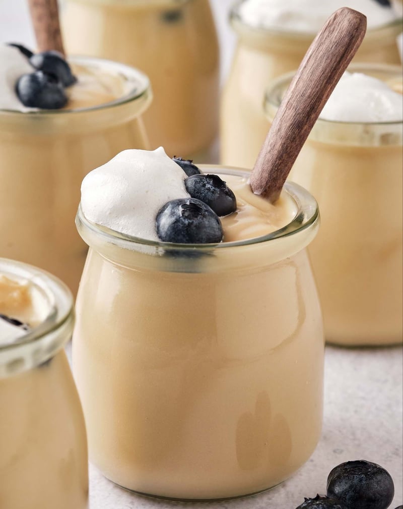 Many jars of vegan vanilla pudding, topped with blueberries and whipped cream, with wooden spoons.