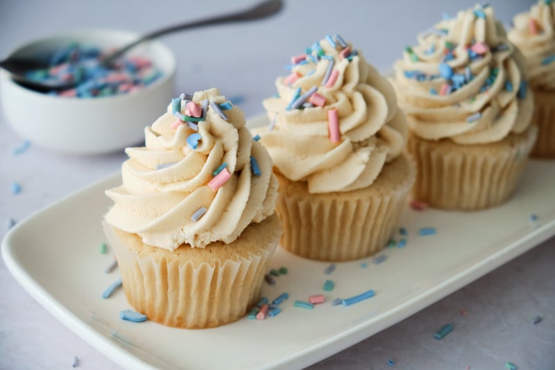 Vegan vanilla cupcakes on a long tray with a bowl of sprinkles in the background.