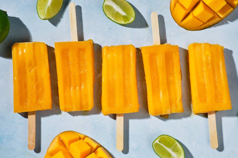 Mango Popsicles on a table, surrounded by lime wedges and sliced mangoes.