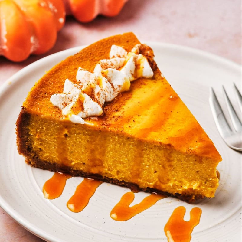 A slice of vegan pumpkin cheesecake topped with vegan whipped cream and caramel, on a plate with a fork; mini pumpkins are in the background.