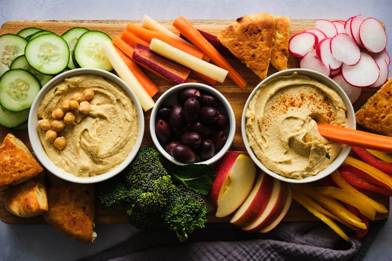 A platter with two bowls of hummus, a bowl of olives, and various vegetables and pita triangles