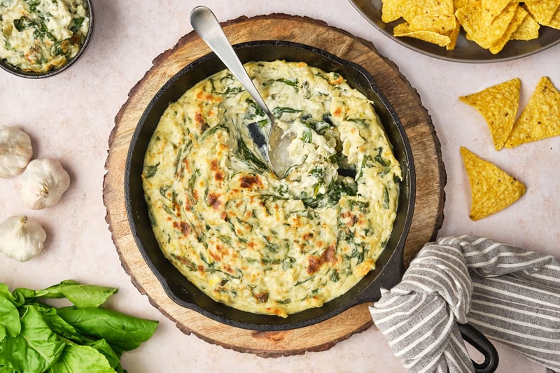 A top-down view of vegan spinach and artichoke dip in a cast-iron pan, with a spoon. To the sides are garlic, spinach, chips, and a bowl of dip.